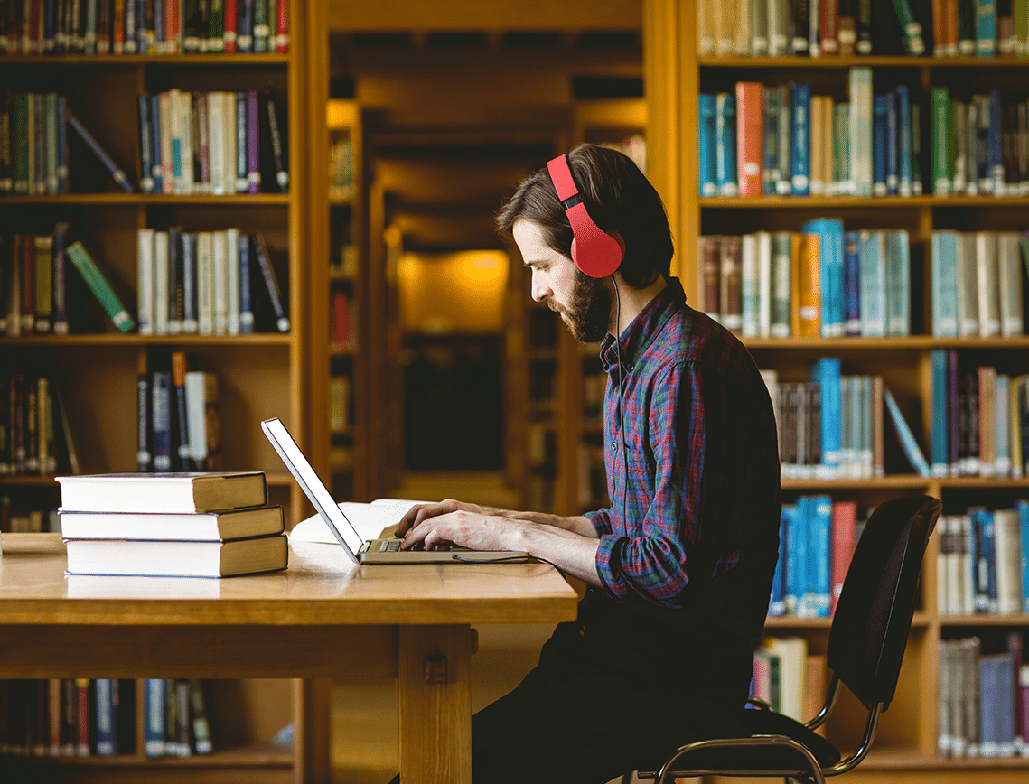 a man works on an assignment in a college library while wearing red headphones